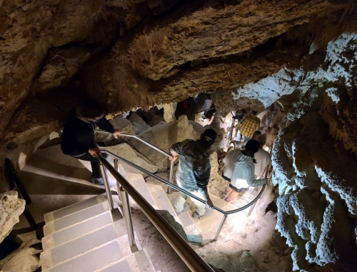 People descend a winding staircase in a cave, surrounded by rocky walls and dim lighting.
