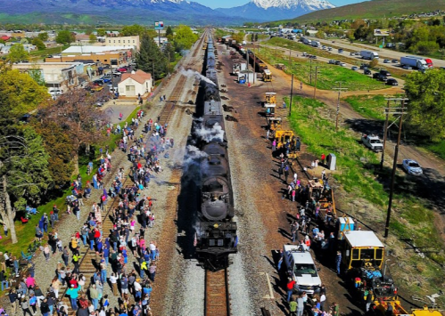 Aerial view of a steam train on tracks, surrounded by a crowd of people and scenic mountains in the background.