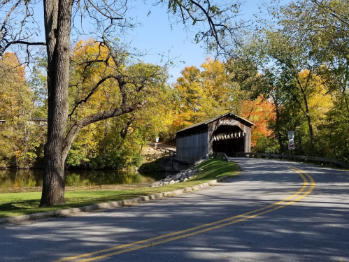 A scenic view of a winding road leading to a covered bridge, surrounded by colorful autumn foliage and a calm river.