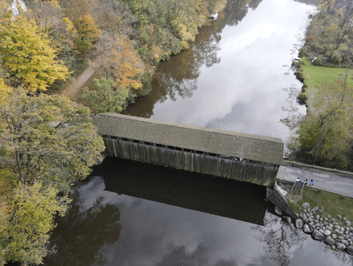 Aerial view of a covered bridge over a calm river, surrounded by autumn foliage and a winding path.