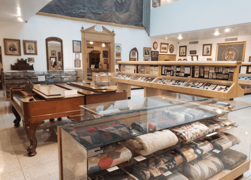 A museum interior featuring display cases with textiles and artifacts, surrounded by framed portraits and historical items.