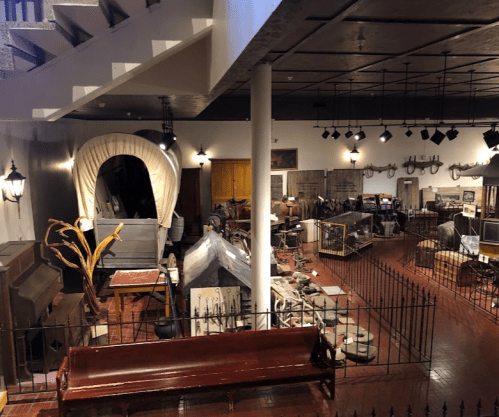 A spacious museum interior filled with historical artifacts, including wagons, furniture, and various displays.