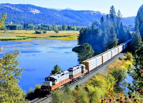 A train travels along a scenic route beside a river, surrounded by trees and mountains in the background.