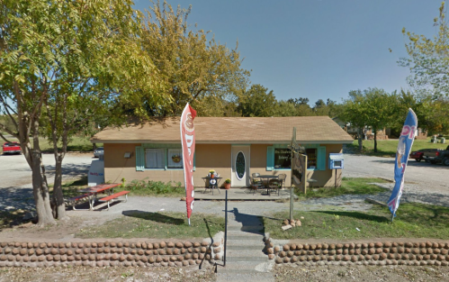 A small, tan building with a front porch, surrounded by trees and flags, in a gravel parking area.