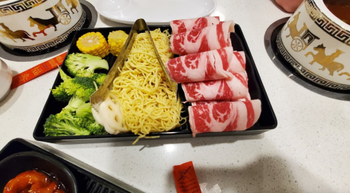 A black tray with broccoli, corn, noodles, and slices of meat, ready for cooking.
