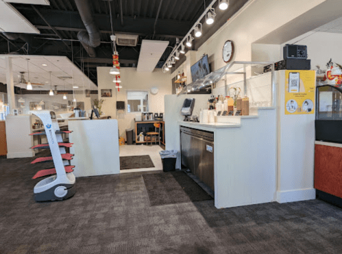 A modern café interior featuring a service counter, seating area, and a robotic assistant on the floor.