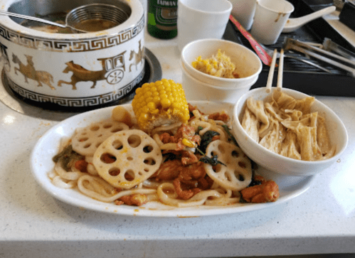 A plate of noodles with chicken, lotus root, and corn, alongside a bowl of rice and a container of soup.