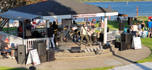 A band performs on stage at a waterfront park, with audience members seated nearby and sound equipment set up.