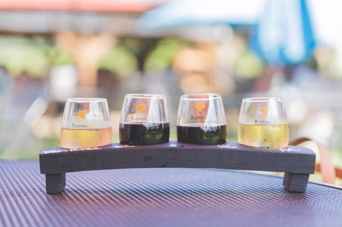 Four glasses of different beverages arranged on a wooden flight board, set on a table outdoors.