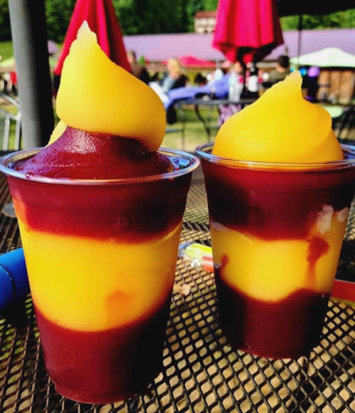 Two cups of layered frozen dessert, one with yellow mango and the other with dark red berry swirl, on a table.