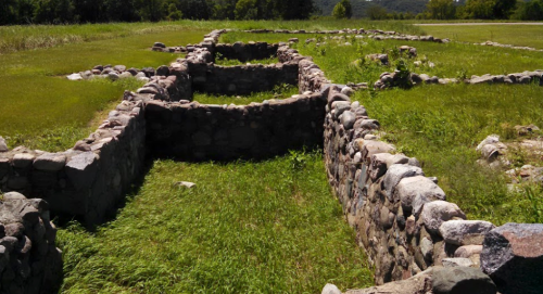 Ruins of stone walls in a grassy area, with trees and hills in the background under a clear blue sky.