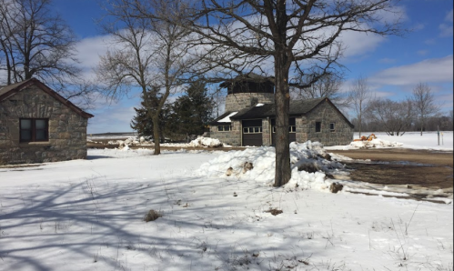 A snowy landscape featuring stone buildings and bare trees under a partly cloudy sky.