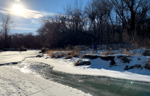 A snowy riverbank with a person standing near the water, surrounded by trees and sunlight.