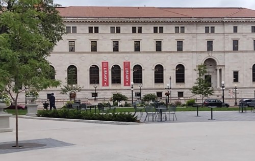 A historic building with large windows and banners, surrounded by greenery and outdoor seating in a public space.