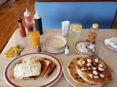 A table with breakfast items: pancakes with butter, eggs on toast, orange juice, and a bowl of grits.