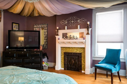 Cozy bedroom featuring a TV, decorative fireplace, and a blue chair under a draped ceiling.