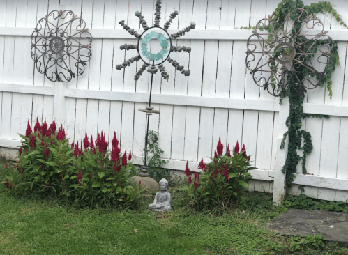 A garden scene featuring red flowers, a small statue, and decorative metal wall art against a white fence.