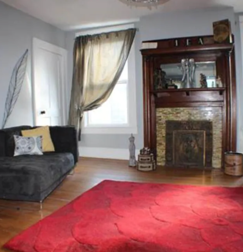 Cozy living room featuring a dark sofa, a red rug, a decorative fireplace, and a large window with drapes.