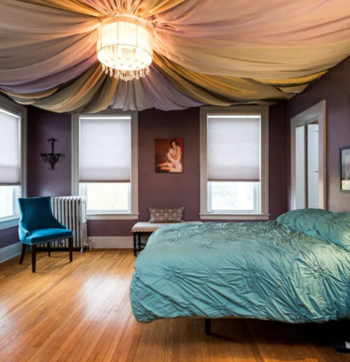 A cozy bedroom with a teal bedspread, a chandelier, and large windows with shades, featuring a chair and artwork.