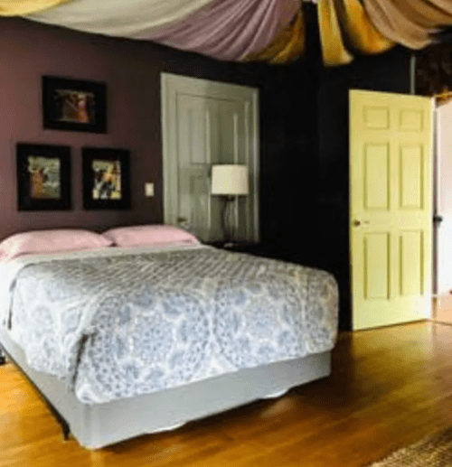Cozy bedroom with a patterned bedspread, soft lighting, and colorful draped ceiling. Walls are dark purple.