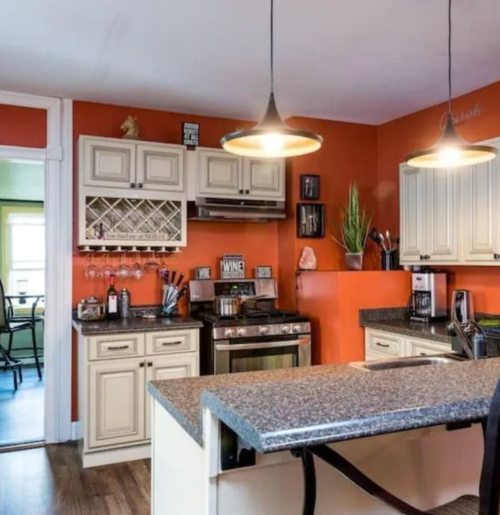 A modern kitchen with orange walls, a granite countertop, and stylish lighting, featuring wine storage and appliances.