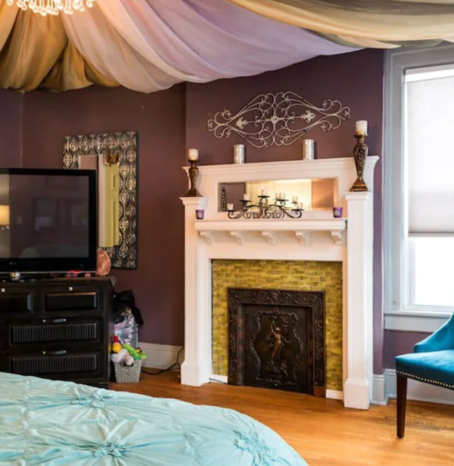 Cozy bedroom with a decorative fireplace, TV, and elegant drapery overhead. Warm colors and stylish decor create a welcoming atmosphere.
