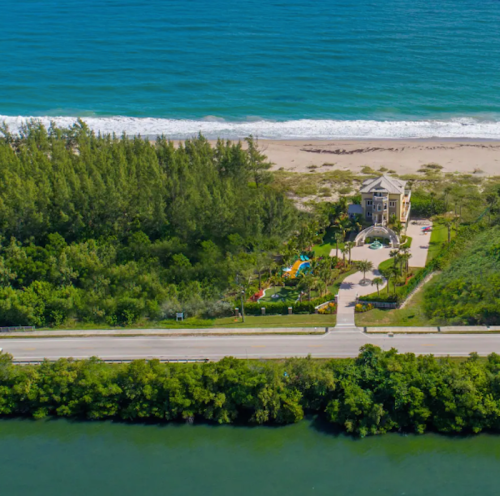 Aerial view of a beachside mansion surrounded by greenery, with a road and ocean in the background.