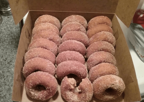 A box filled with freshly made, sugar-coated donuts arranged in rows. One donut is partially eaten.