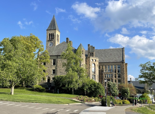 Historic stone building with a clock tower, surrounded by green trees and a blue sky with scattered clouds.