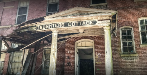Weathered entrance of "Daughters Cottage" with peeling paint and a dilapidated porch, set against a brick building.