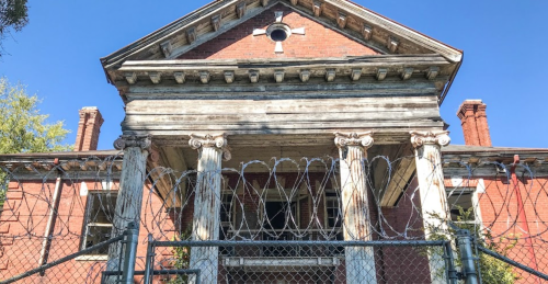 Abandoned building with a classical facade, surrounded by a barbed wire fence under a clear blue sky.