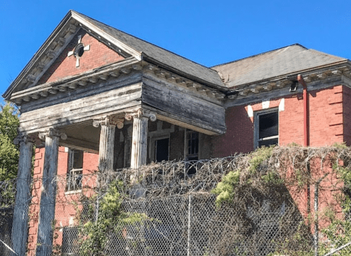 Abandoned brick building with classical columns, overgrown vegetation, and a chain-link fence under a clear blue sky.