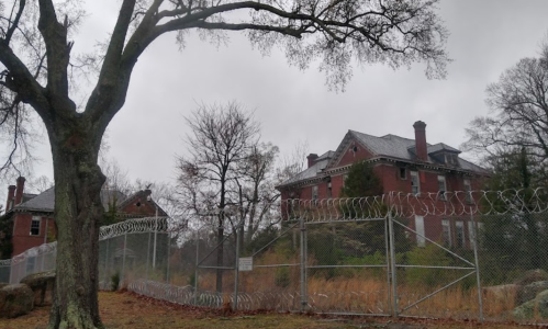A dilapidated brick mansion surrounded by a barbed wire fence, with overcast skies and bare trees.