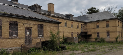 Abandoned yellow brick building with boarded windows, overgrown grass, and a cloudy sky.