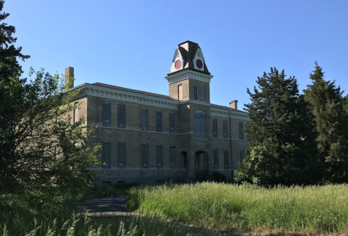 Abandoned brick building with a distinctive tower, surrounded by tall grass and trees under a clear blue sky.