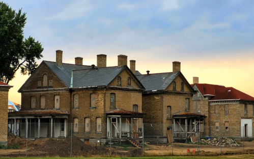 Abandoned brick buildings with multiple chimneys, surrounded by construction debris and a cloudy sky.