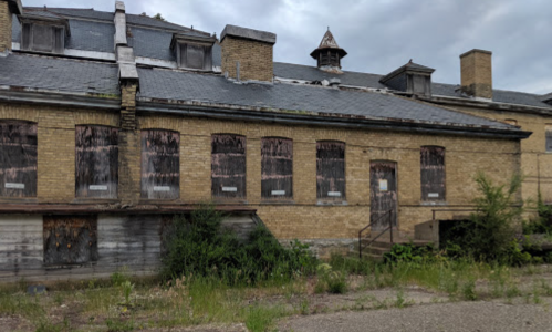 Abandoned brick building with boarded windows, overgrown grass, and a cloudy sky in the background.