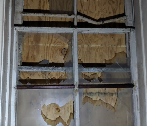 Broken window with shattered glass and wooden boards covering the gaps, creating a distressed, abandoned look.