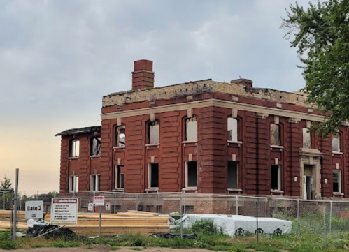 Abandoned brick building with broken windows, surrounded by construction materials and a fence. Overcast sky in the background.