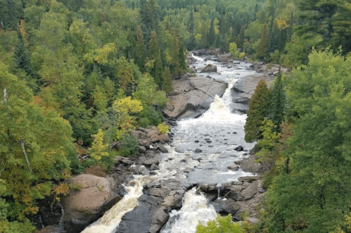 A scenic view of a river flowing through rocky terrain, surrounded by lush green trees and autumn foliage.