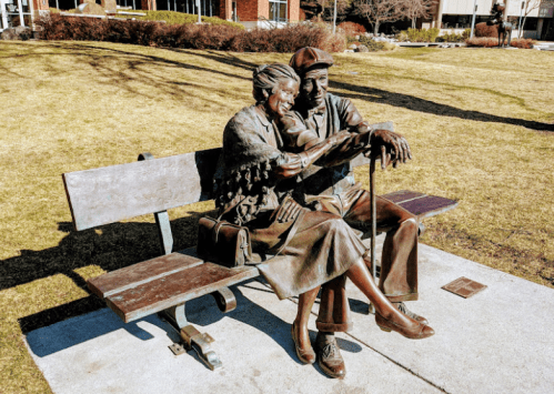 A bronze sculpture of an elderly couple sitting closely on a bench, sharing a moment together in a park.