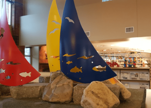 Colorful sail-shaped sculptures in red, yellow, and blue, surrounded by rocks, inside a bright indoor space.