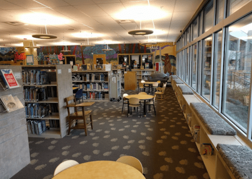 A spacious library interior with bookshelves, tables, and seating areas, featuring large windows and colorful wall art.