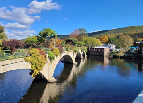A scenic view of a stone bridge over a calm river, surrounded by trees and colorful autumn foliage under a blue sky.
