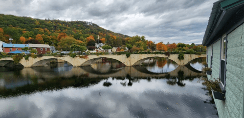A scenic view of a stone bridge over a calm river, surrounded by autumn foliage and cloudy skies.