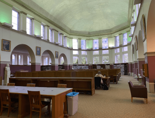 A spacious library interior with tall windows, wooden tables, and a person studying at a desk.