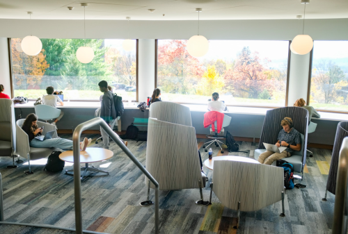 A cozy study area with students reading and working by large windows, surrounded by autumn foliage outside.