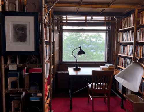 Cozy library corner with bookshelves, a desk, chair, and a window overlooking greenery. Warm, inviting atmosphere.