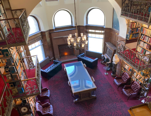 A spacious library with high ceilings, large windows, elegant furniture, and a central table surrounded by bookshelves.