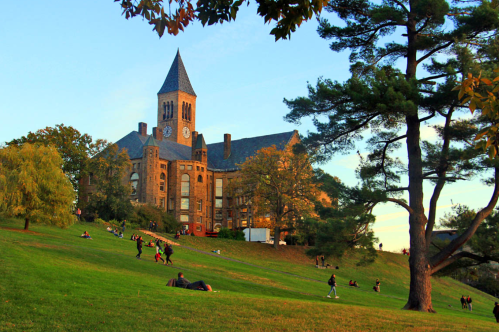 A historic building with a clock tower overlooks a grassy hill where people relax and enjoy the outdoors at sunset.